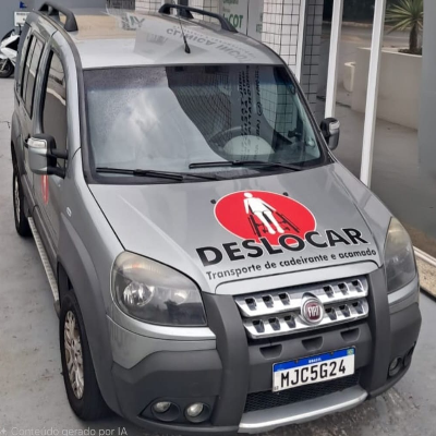 A foto é de um carro utilitário cinza estacionado em frente a um prédio com fachada de vidro. O veículo é visto de frente em um leve ângulo lateral. No capô há um grande adesivo circular vermelho com o símbolo de uma pessoa em cadeira de rodas em branco. Abaixo do símbolo está escrito em letras grandes “DESLOCAR” e, em letras menores, “Transporte de cadeirante e acamado”.